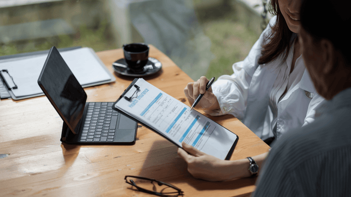 Eine Person, die mit einem Arzt oder Berater über eine Gesundheitsversicherung spricht und ein Formular mit der Aufschrift "Health Insurance" ausfüllt. Auf dem Tisch stehen ein Laptop und eine Kaffeetasse.